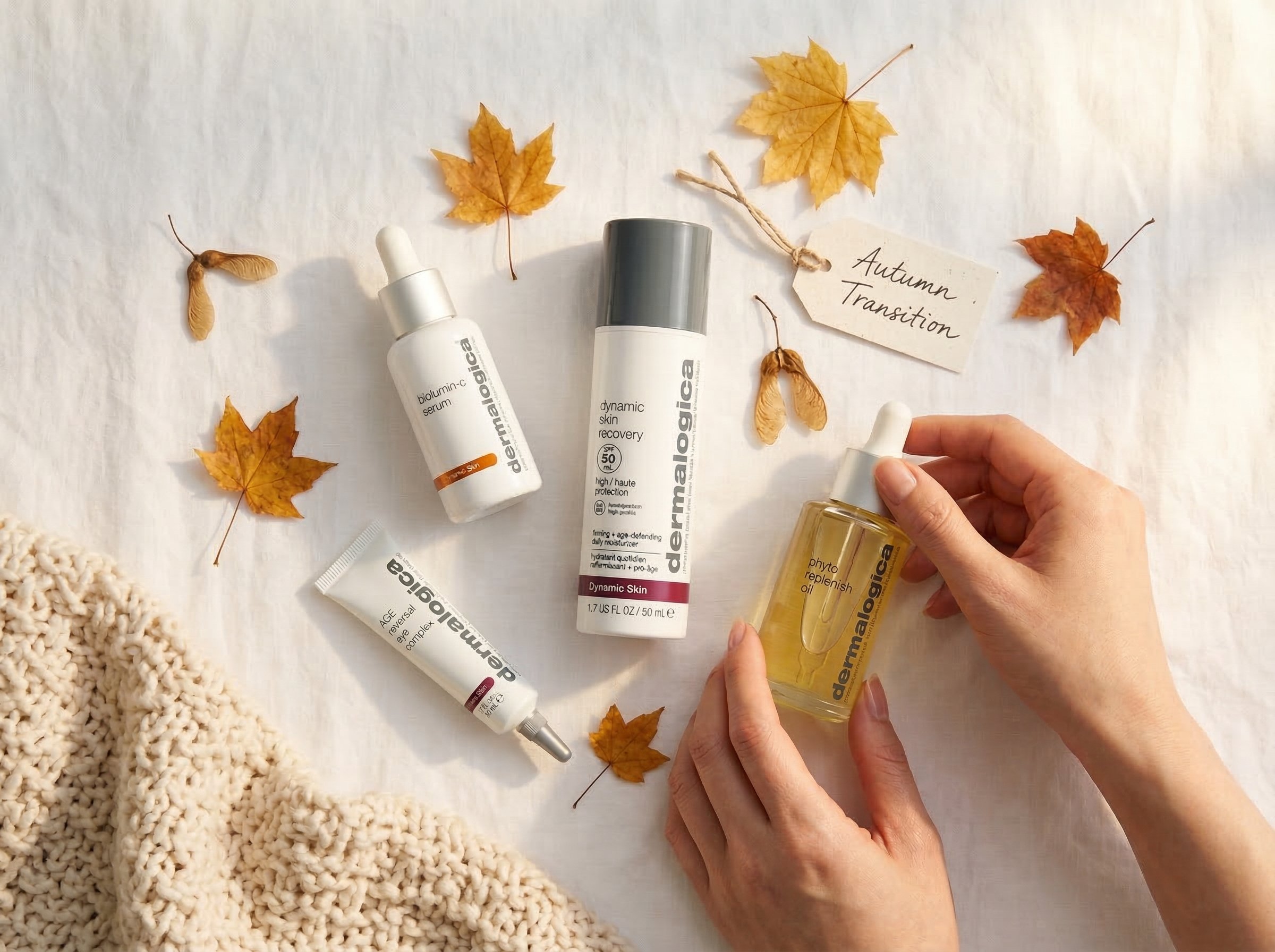 Overhead flat lay of Dermalogica autumn skincare products including vitamin C serum, face oil, eye complex, and SPF arranged with autumn leaves and knit blanket on white surface