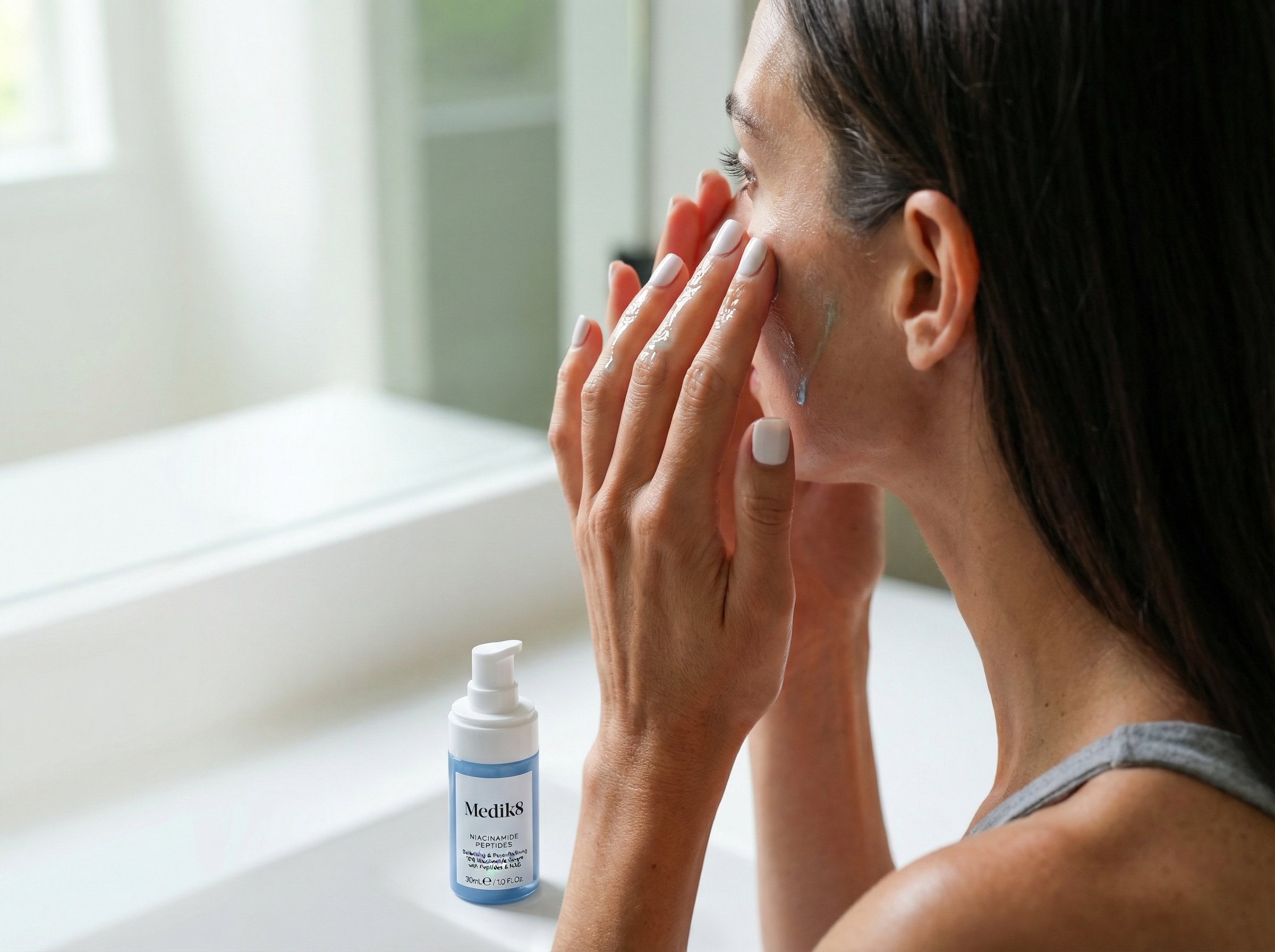 Woman applying niacinamide serum to face with serum bottle on bathroom counter in natural lighting