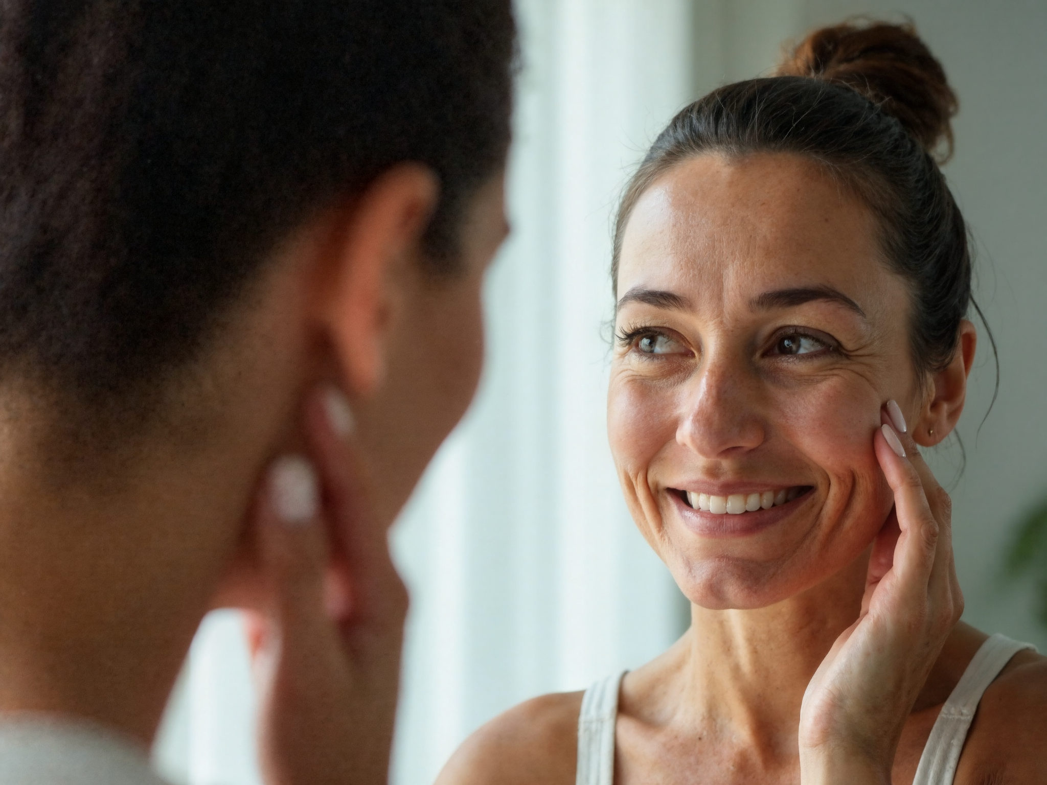 Woman in her early forties looking at her skin in mirror with natural lighting, demonstrating skin assessment and maintenance skincare approach