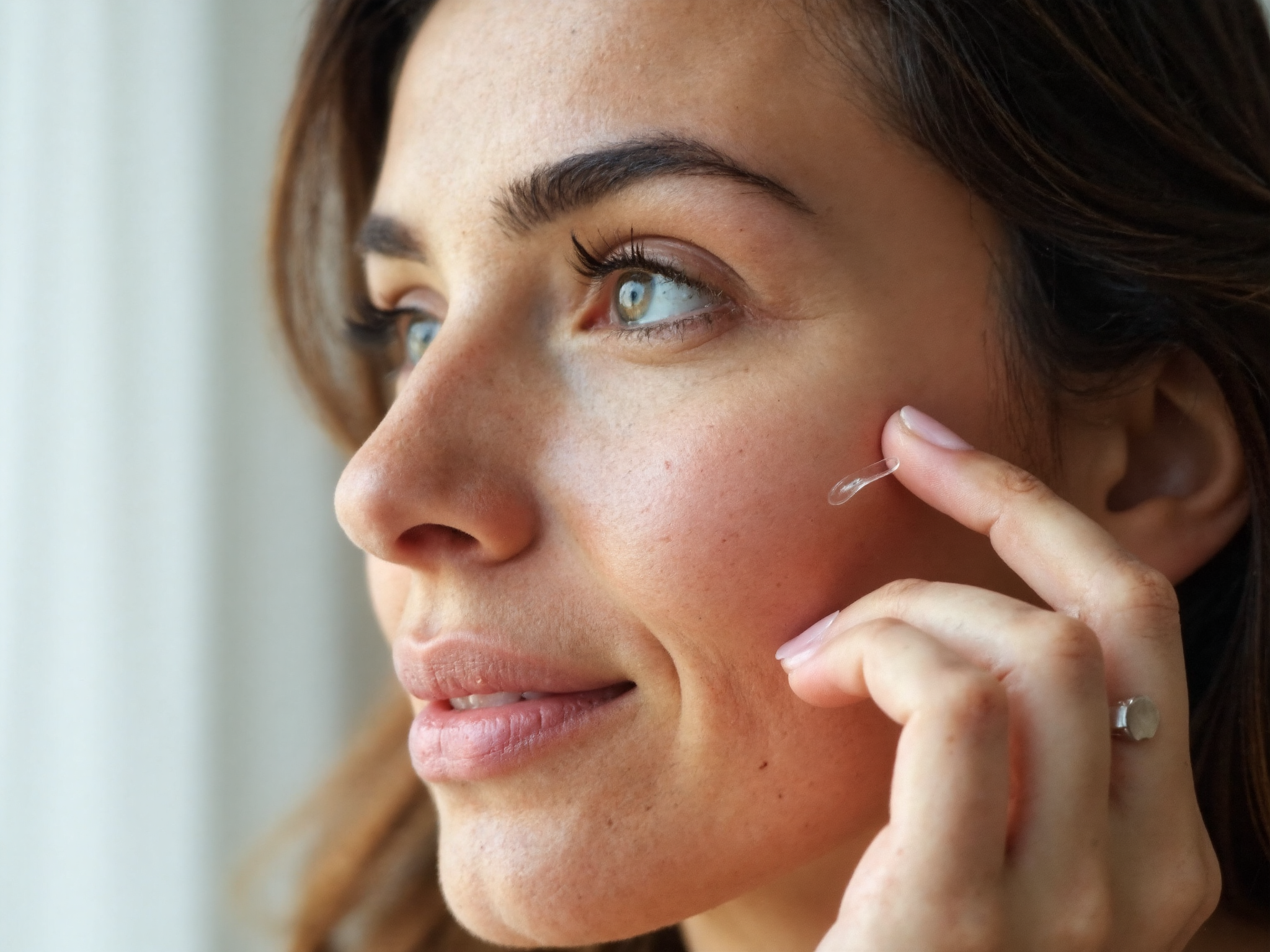 Woman applying clear serum to her cheek with fingertips in natural bathroom lighting showing proper skincare layering technique