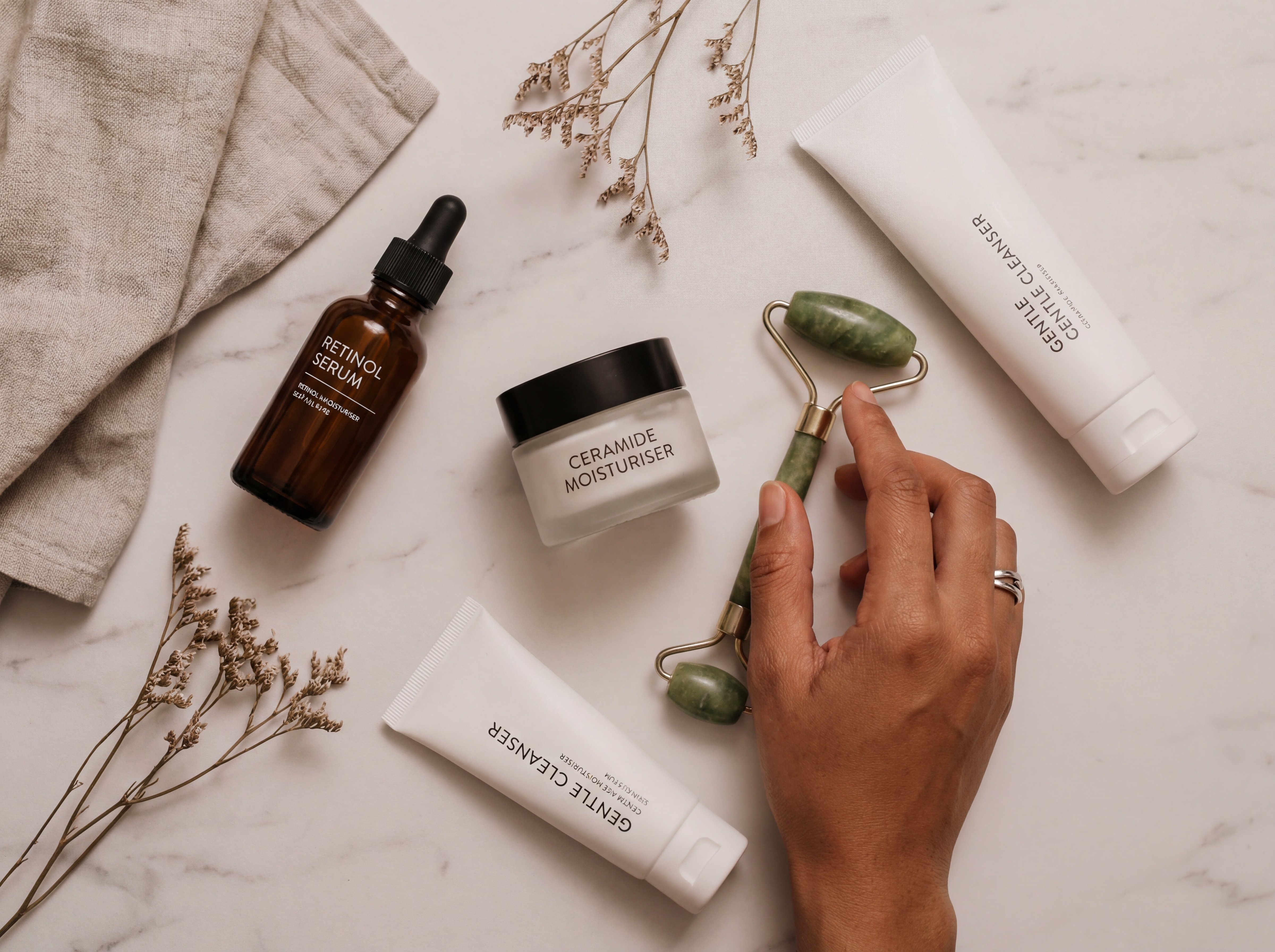Overhead flat lay showing an evening skincare routine on a white marble surface including a retinol serum, ceramide moisturiser, gentle cleanser, and jade roller arranged in a clean editorial style