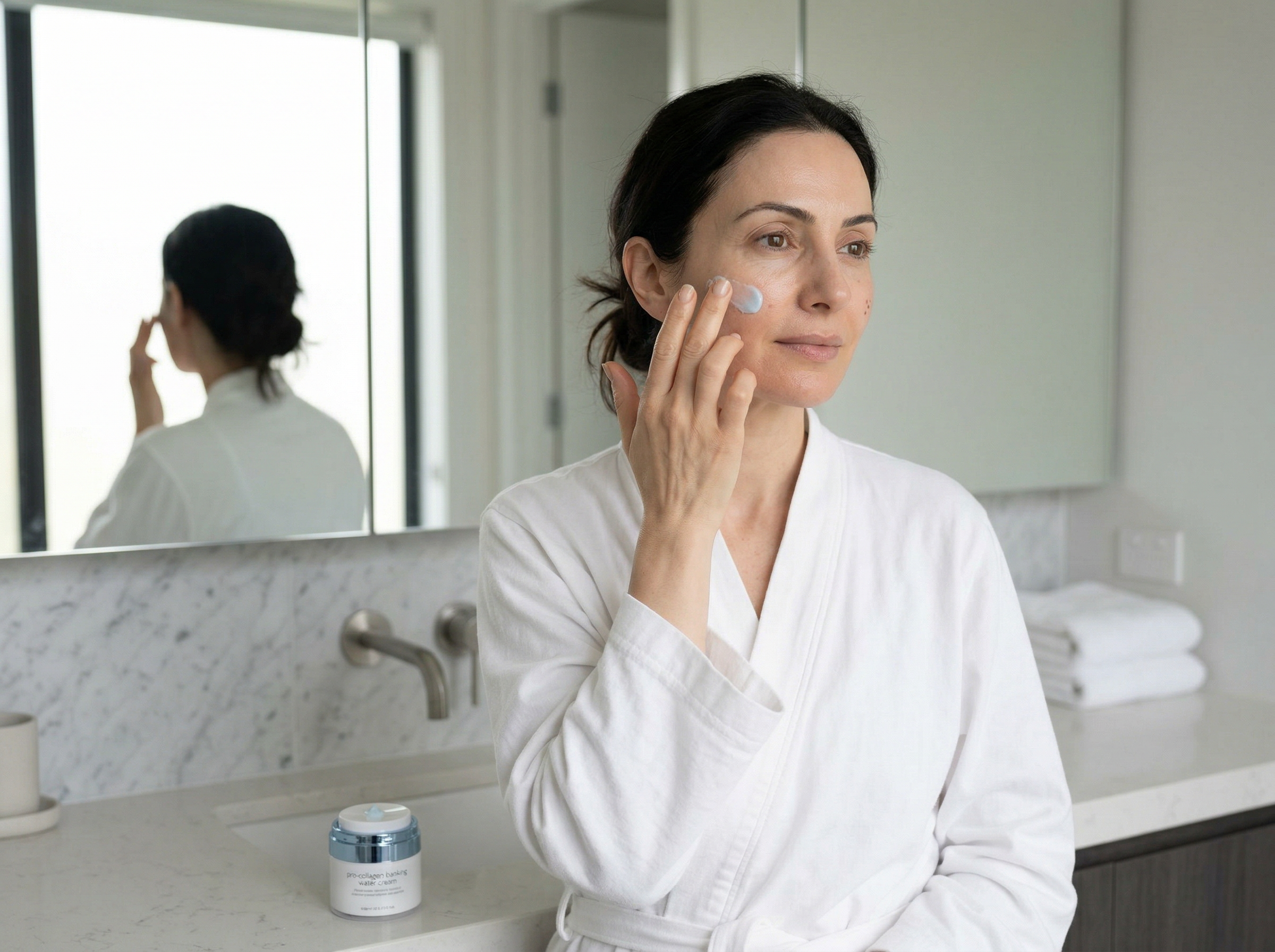 Woman in her 40s applying facial cream to her cheek in a bright, modern bathroom
