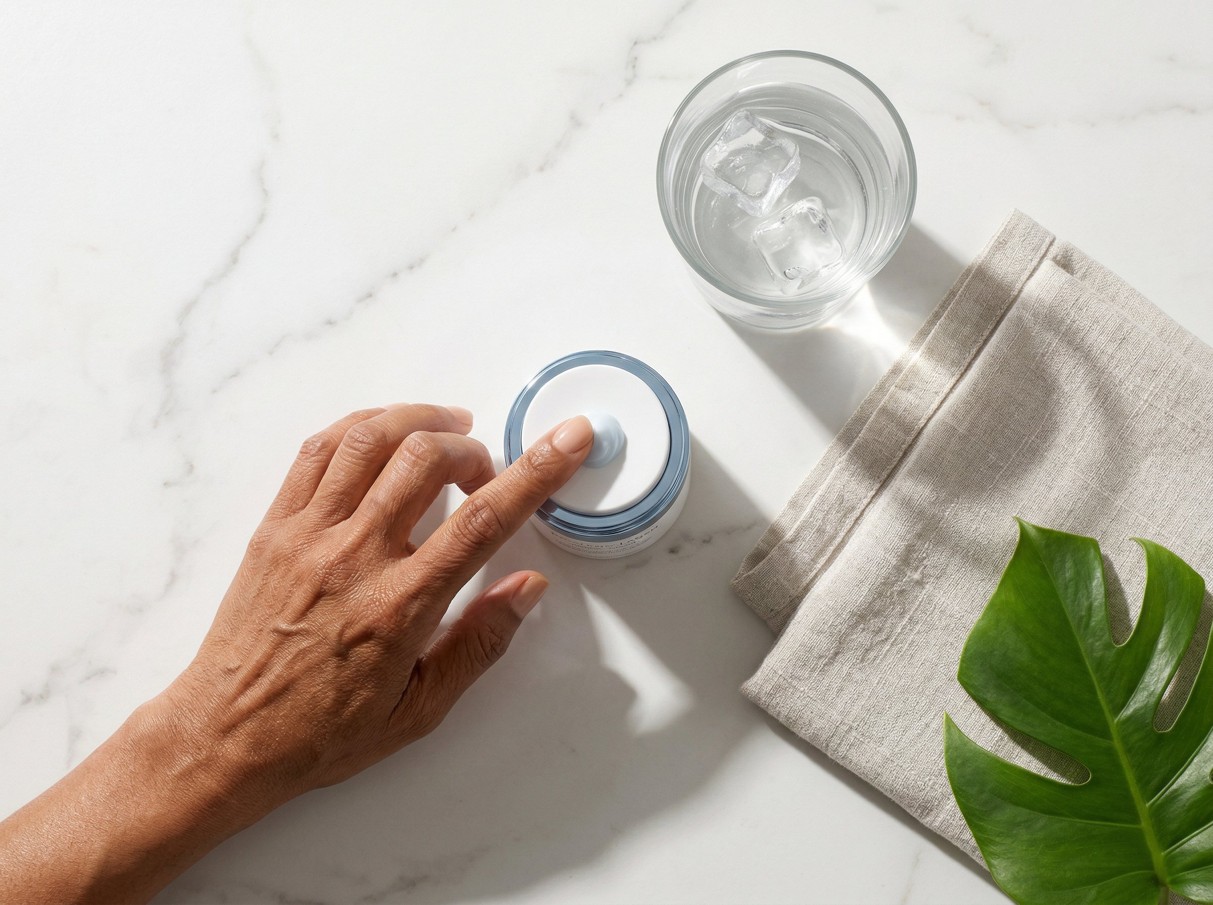 Flat lay of skincare products including niacinamide serum and moisturiser with glass of water on marble surface
