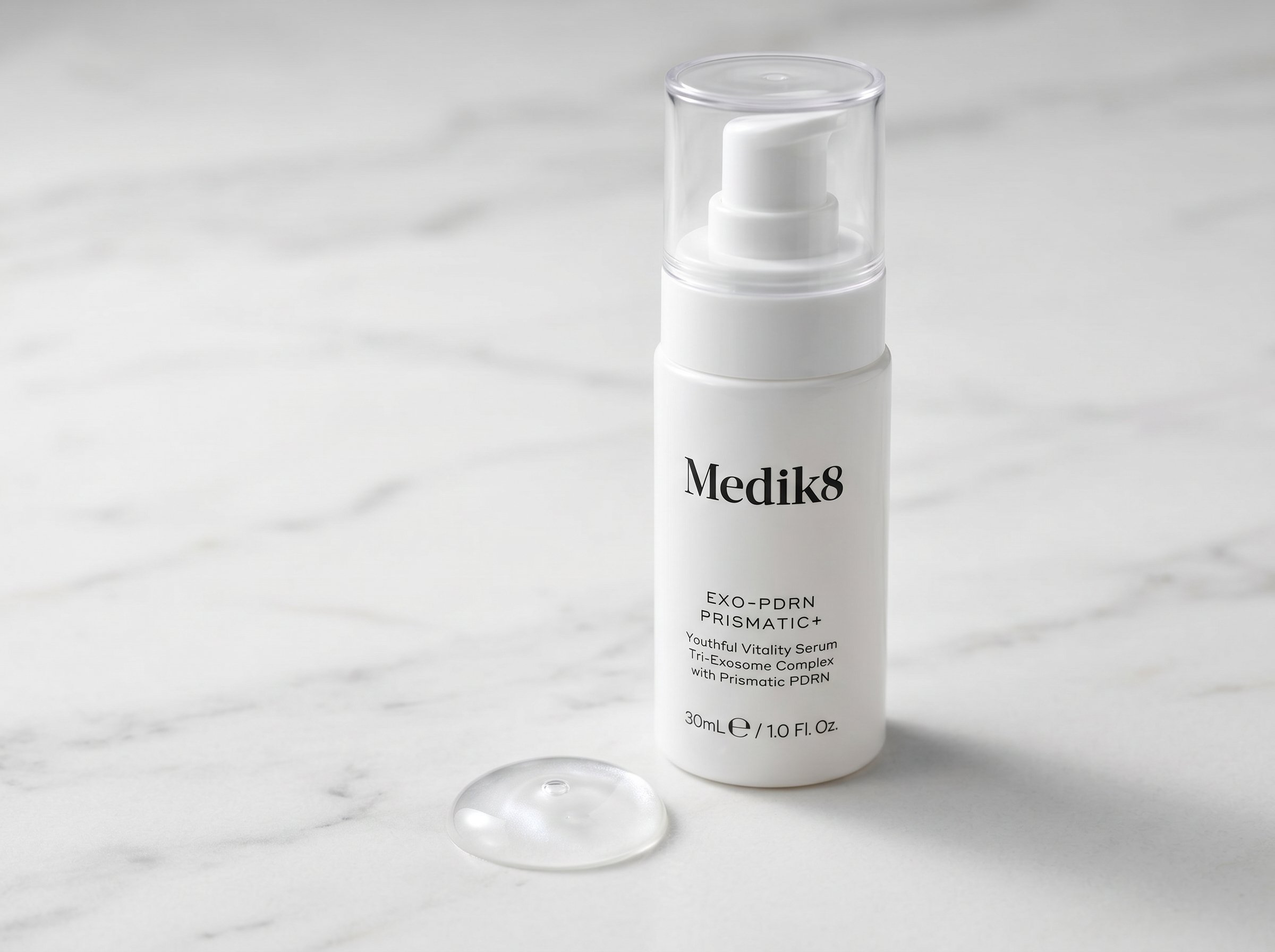 Close-up of Exo-PDRN Prismatic+ serum bottle with a drop of serum visible at the dropper tip, on a white marble surface