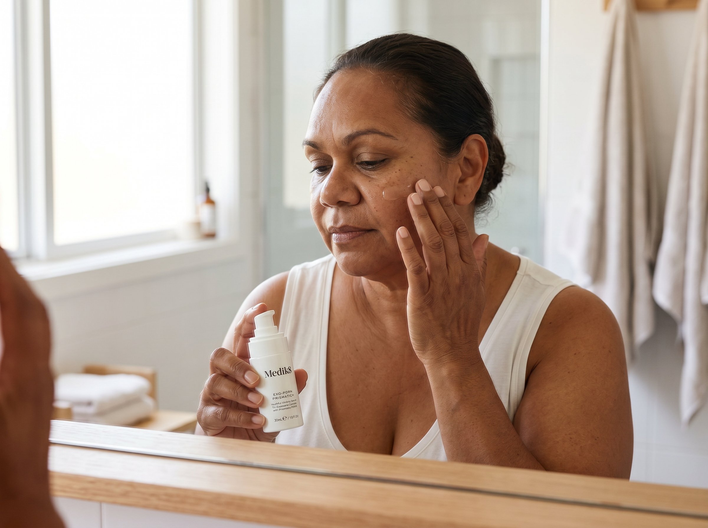 58-year-old Indigenous Australian woman with olive skin applying serum to her cheek in a bright bathroom with natural light