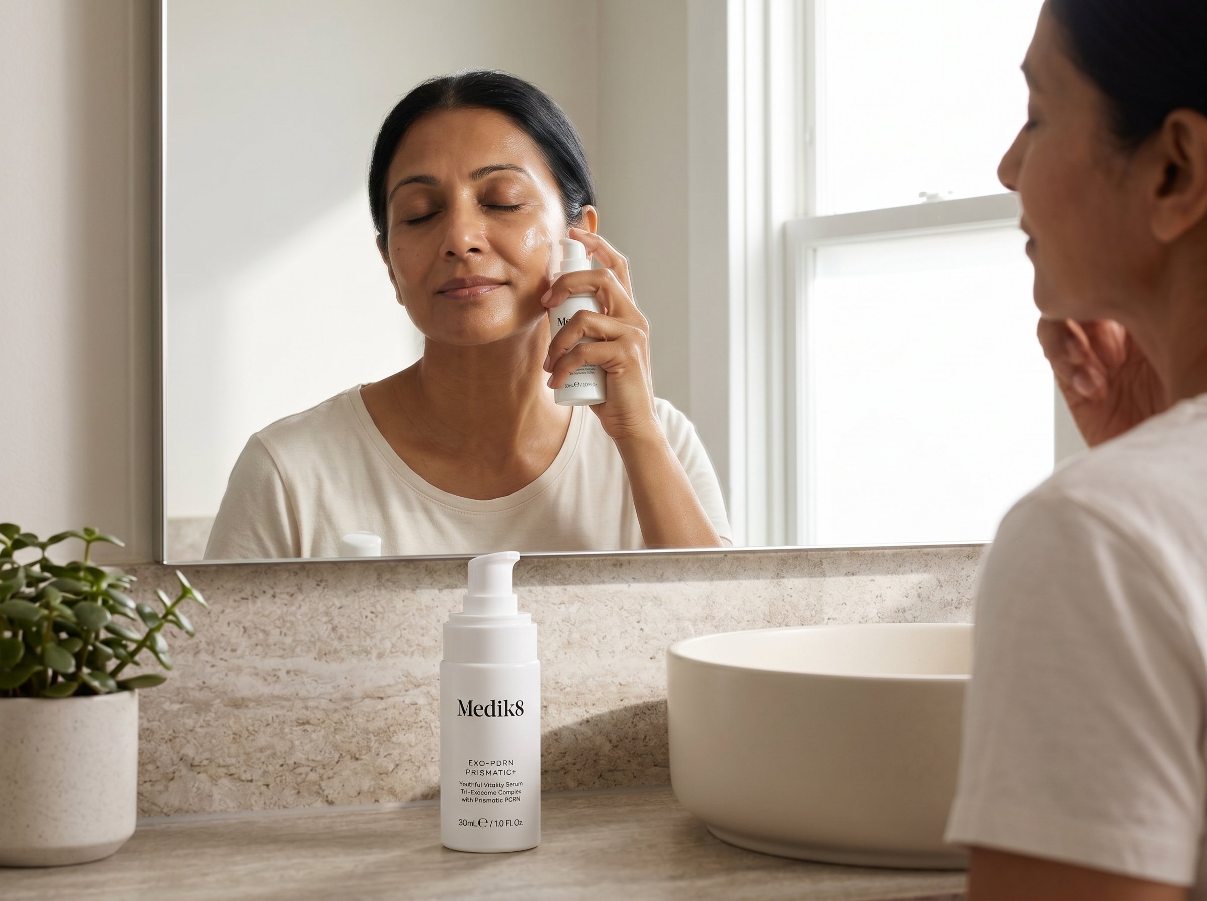 54-year-old South Asian Australian woman with olive skin applying serum to her cheek in a bright bathroom with natural morning light